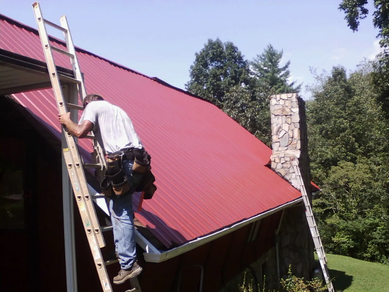 Roofer on a ladder beside a red metal roof and stone chimney — Soffit & Fascia Repair by Roofing Brothers in Pine Bluffs